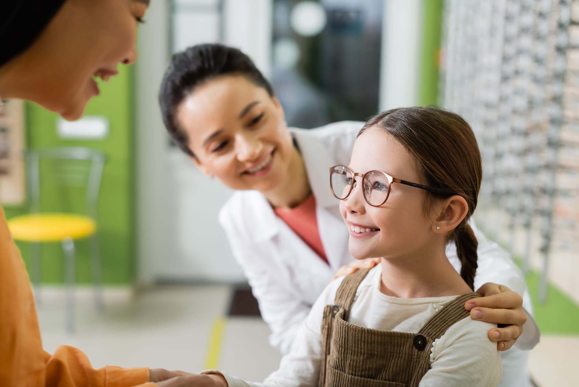 An optometrist and a parent smiling with a young girl trying on new glasses at Vine Eye Care