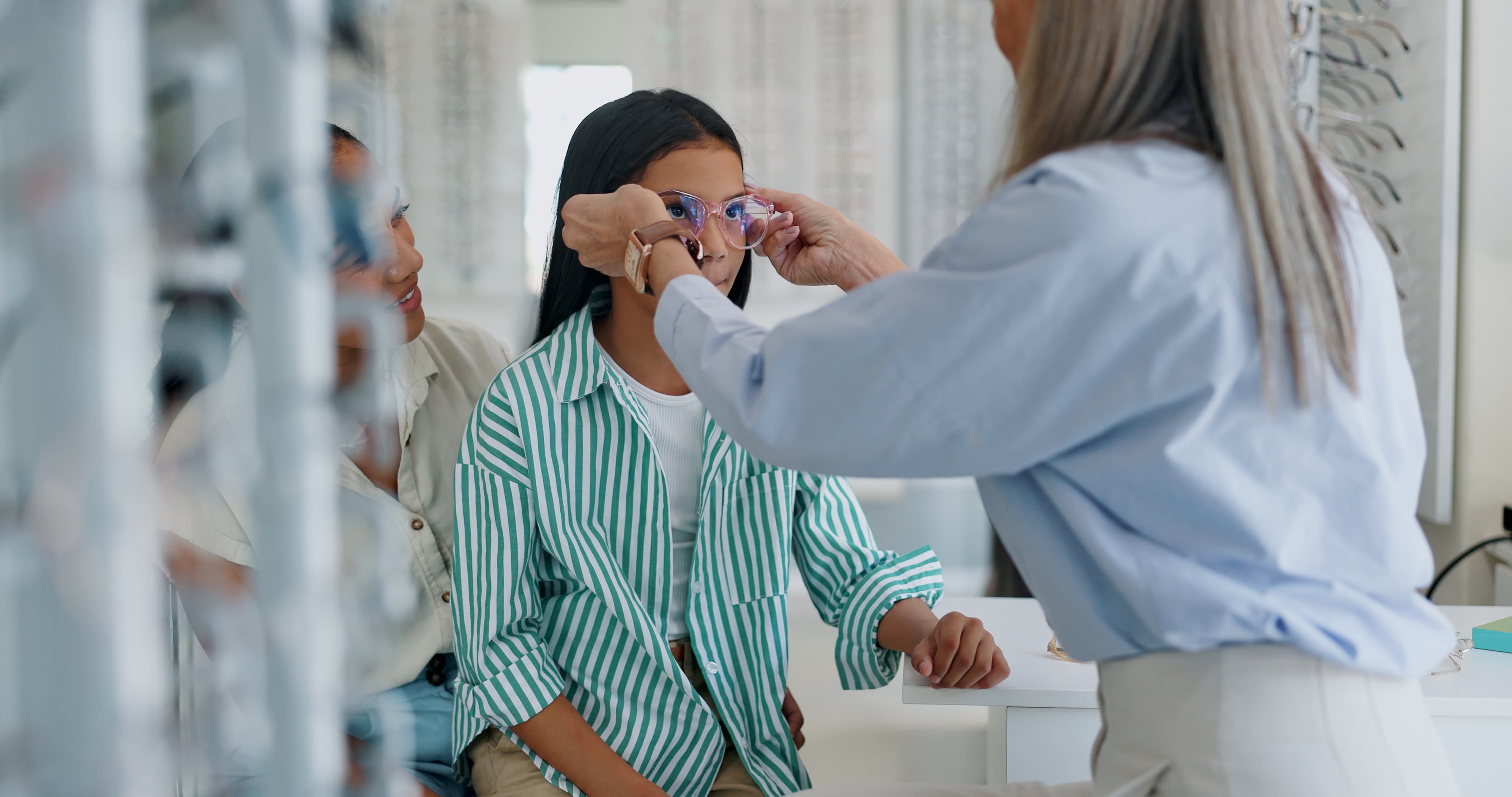 An optometrist fitting glasses on a young patient at Vine Eye Care, with her mother looking on